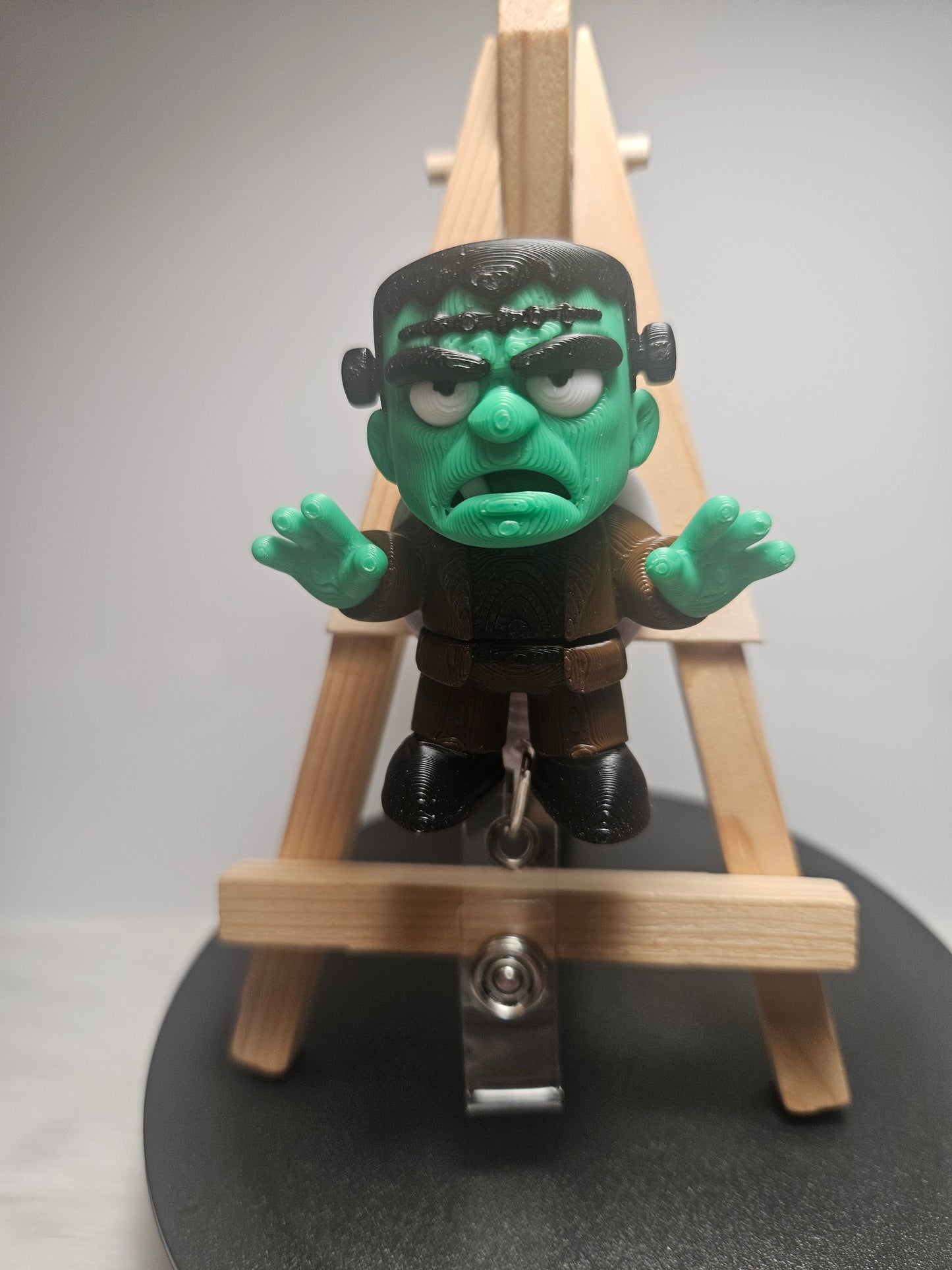A small Frankenstein's monster badgereel with green skin and black hair, posed in front of a miniature wooden easel on a round black display base.