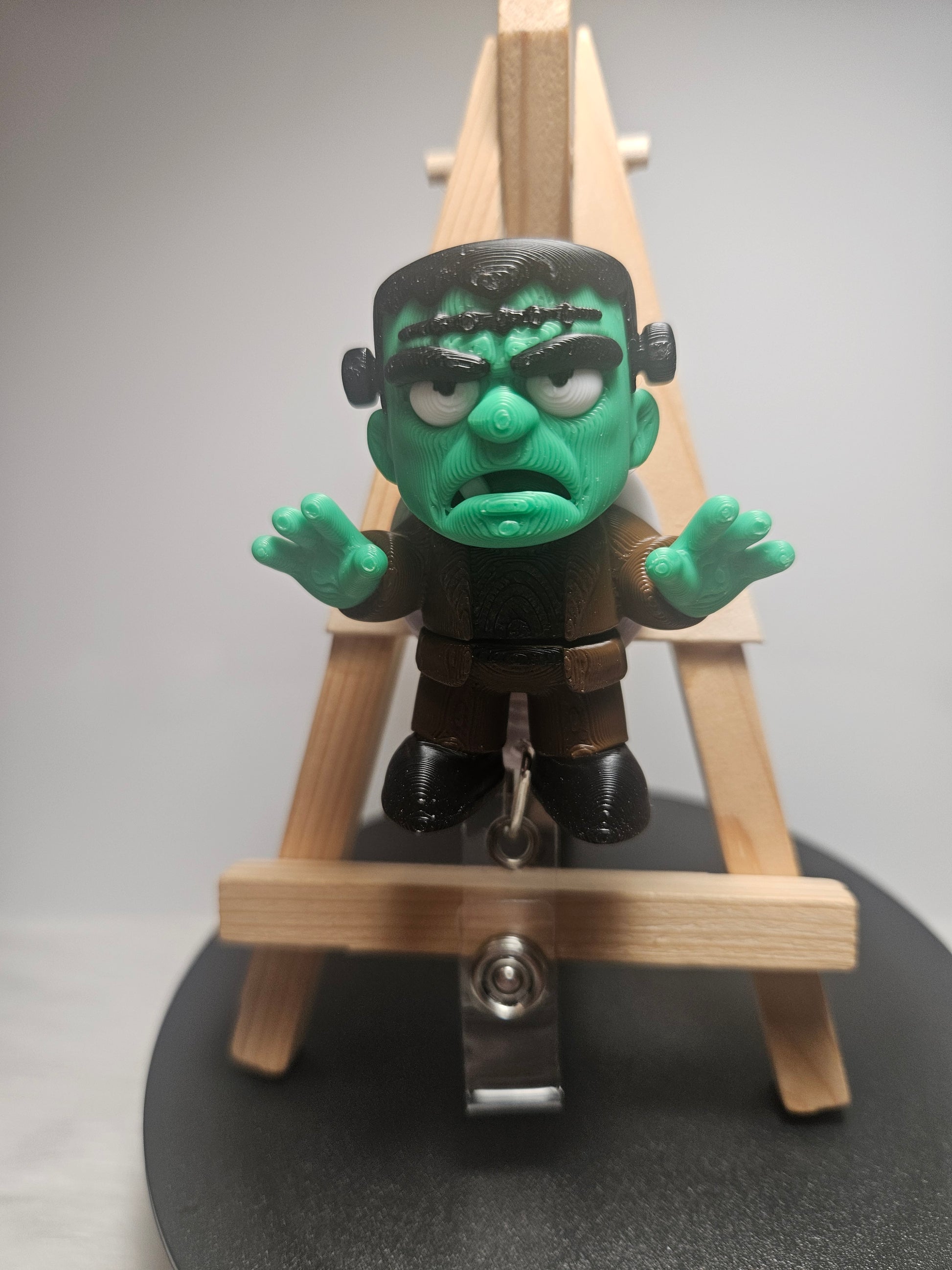 A small Frankenstein's monster badgereel with green skin and black hair, posed in front of a miniature wooden easel on a round black display base.