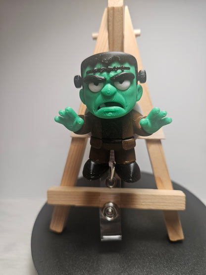 A small Frankenstein's monster badgereel with green skin and black hair, posed in front of a miniature wooden easel on a round black display base.