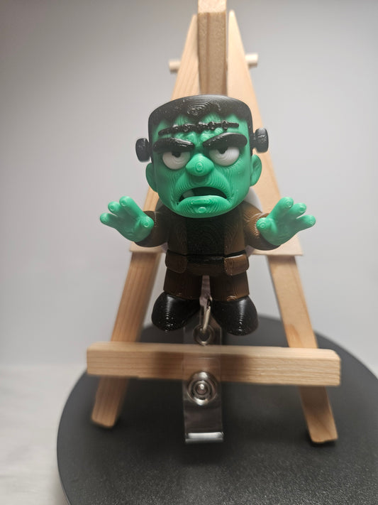A small Frankenstein's monster badgereel with green skin and black hair, posed in front of a miniature wooden easel on a round black display base.