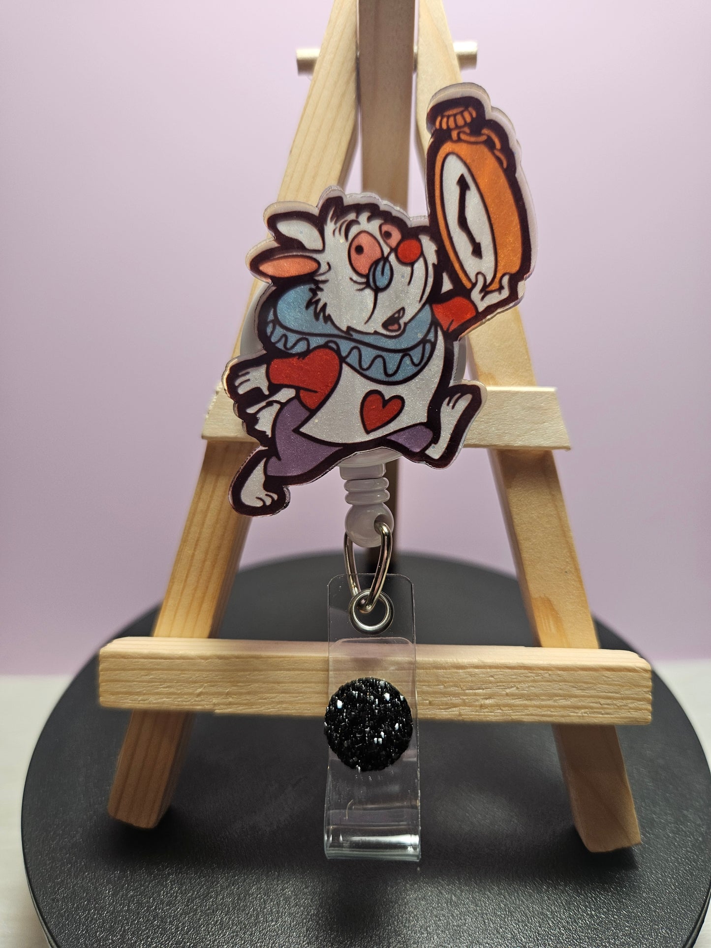 A retractable badge reel featuring an illustration of a character resembling the White Rabbit from Alice in Wonderland, attached to a clear acrylic topper, presented on a wooden display stand.