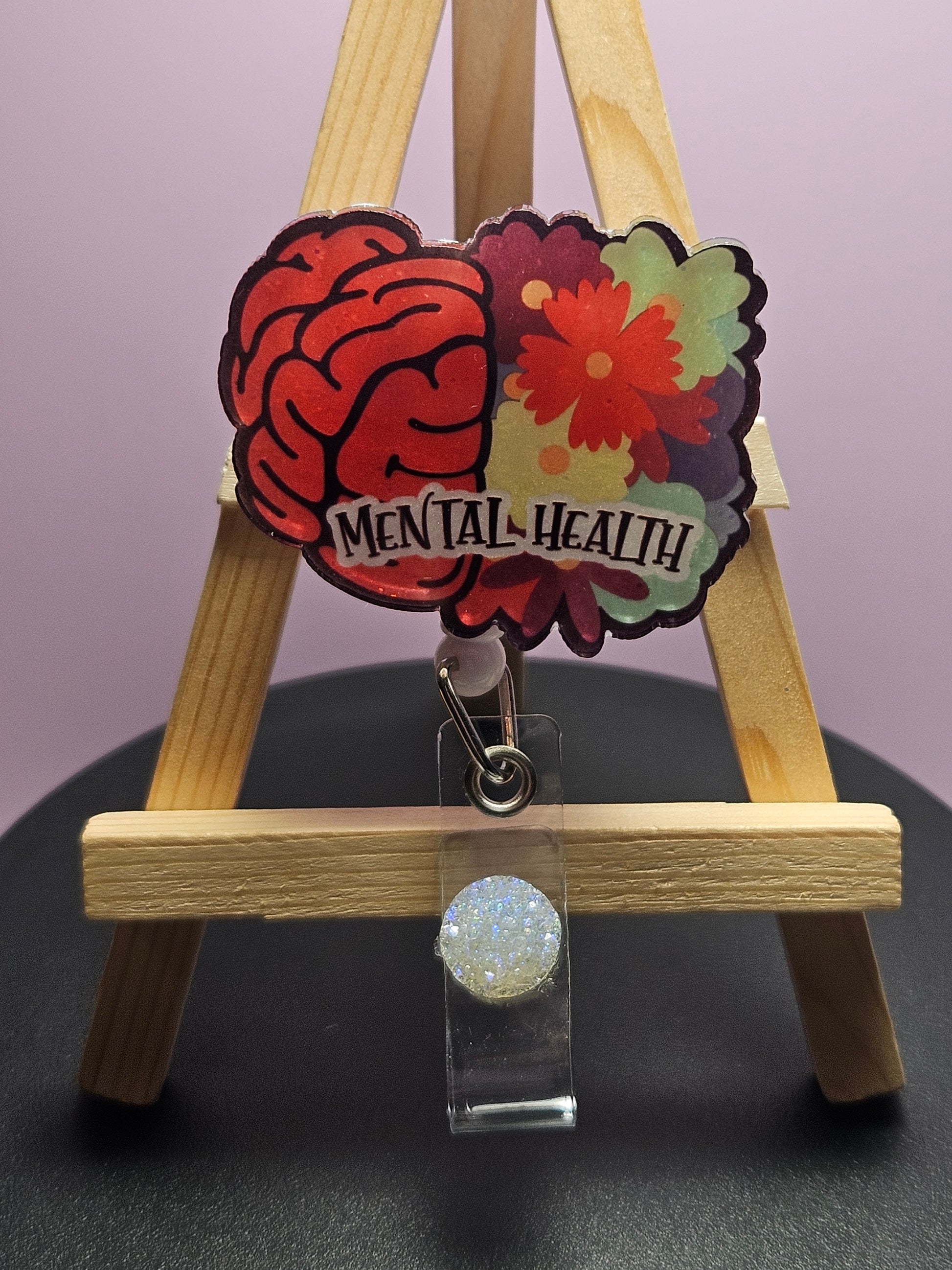 A retractable badge reel with a floral and brain design, labeled 'MENTAL HEALTH', displayed on a wooden easel.