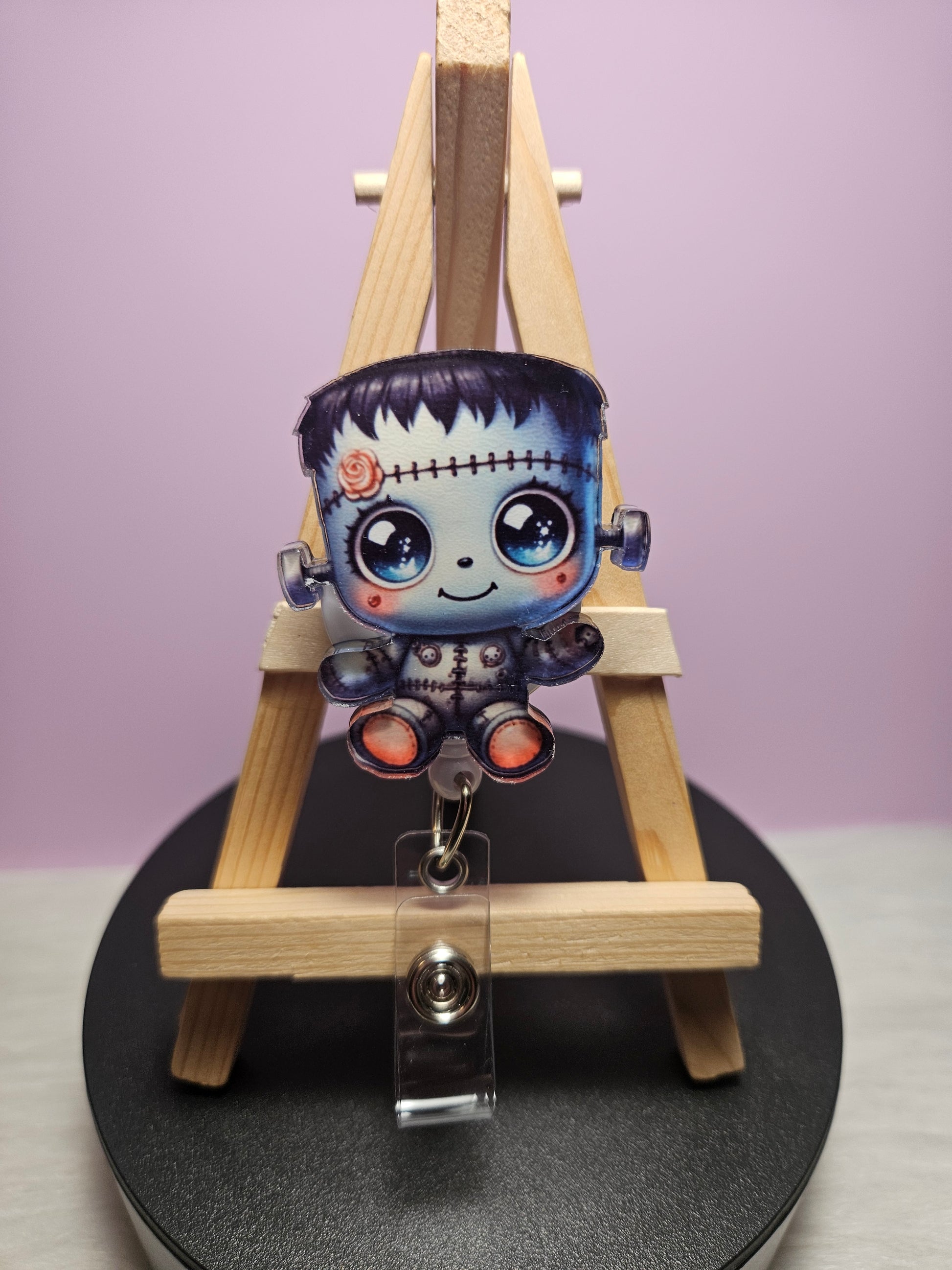 A retractable badge reel featuring a colorful character resembling a cute Frankenstein monster, displayed on a wooden easel.