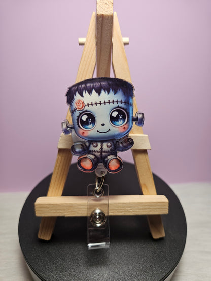 A retractable badge reel featuring a colorful character resembling a cute Frankenstein monster, displayed on a wooden easel.
