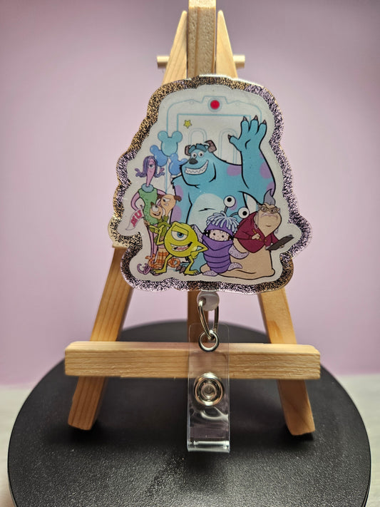 A retractable badge reel featuring a colorful design with animated monster characters displayed on an acrylic topper attached to a wooden easel.