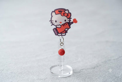 HK badge reel with Hello Kitty face and red microphone charm, hanging on clear stand